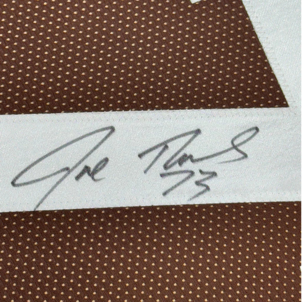 White fabric label with two black ink signatures on brown polka dotted background of framed joe thomas football jersey