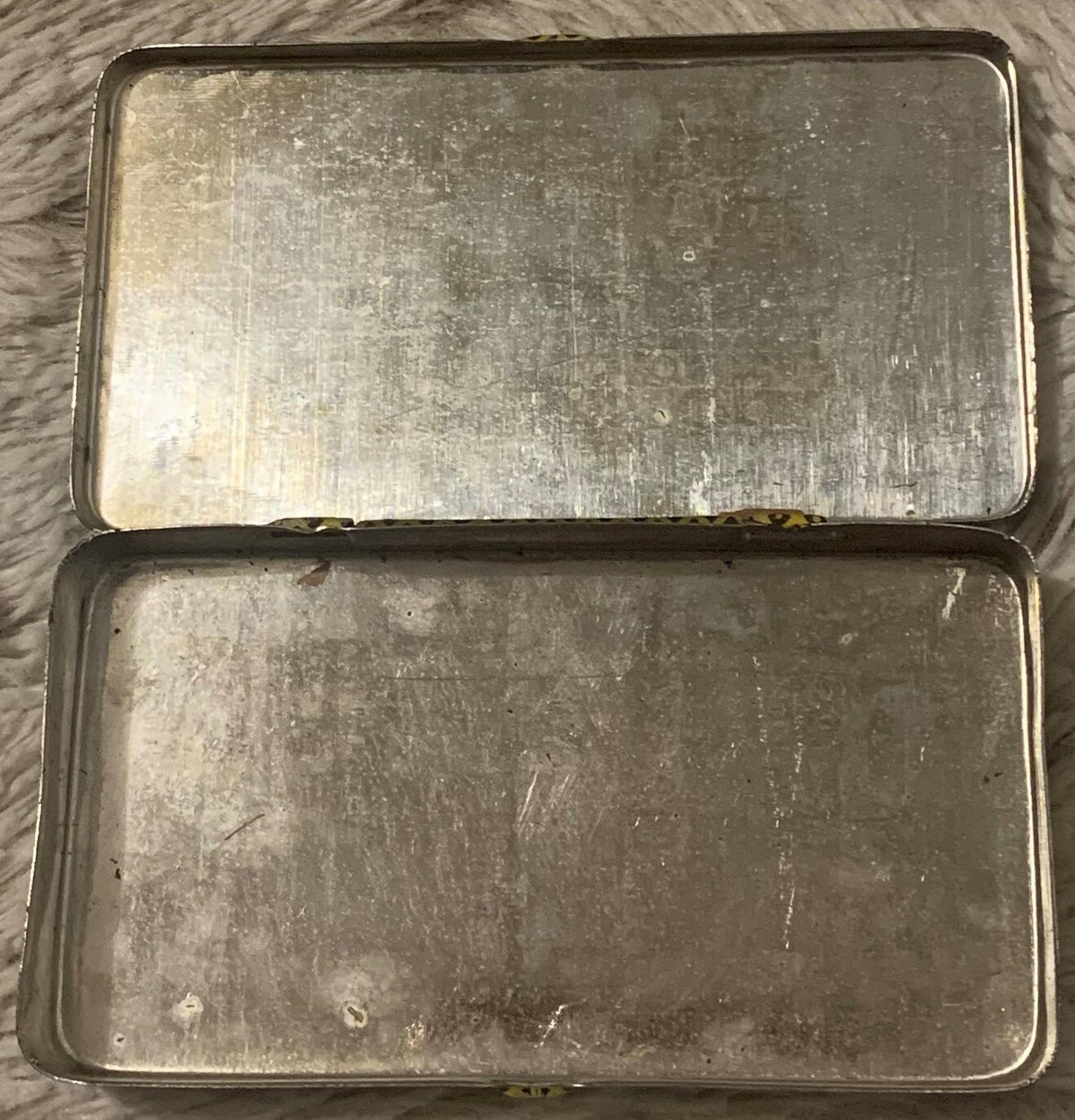 Two vintage metal tins stacked vertically on gray fabric, showing worn silver surfaces with patina and surface scratches