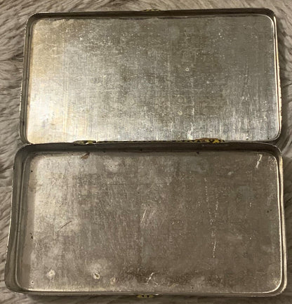 Two vintage metal tins stacked vertically on gray fabric, showing worn silver surfaces with patina and surface scratches
