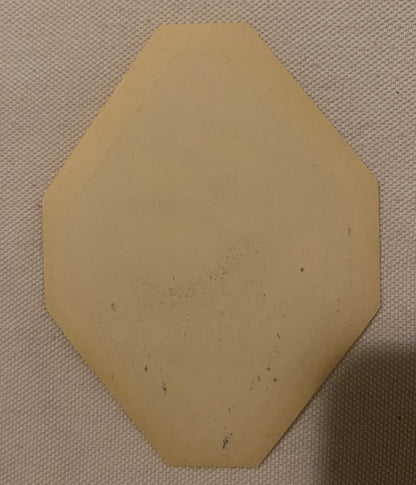 Tan hexagonal paper label with beveled corners on cream fabric background, showing age spots and discoloration
