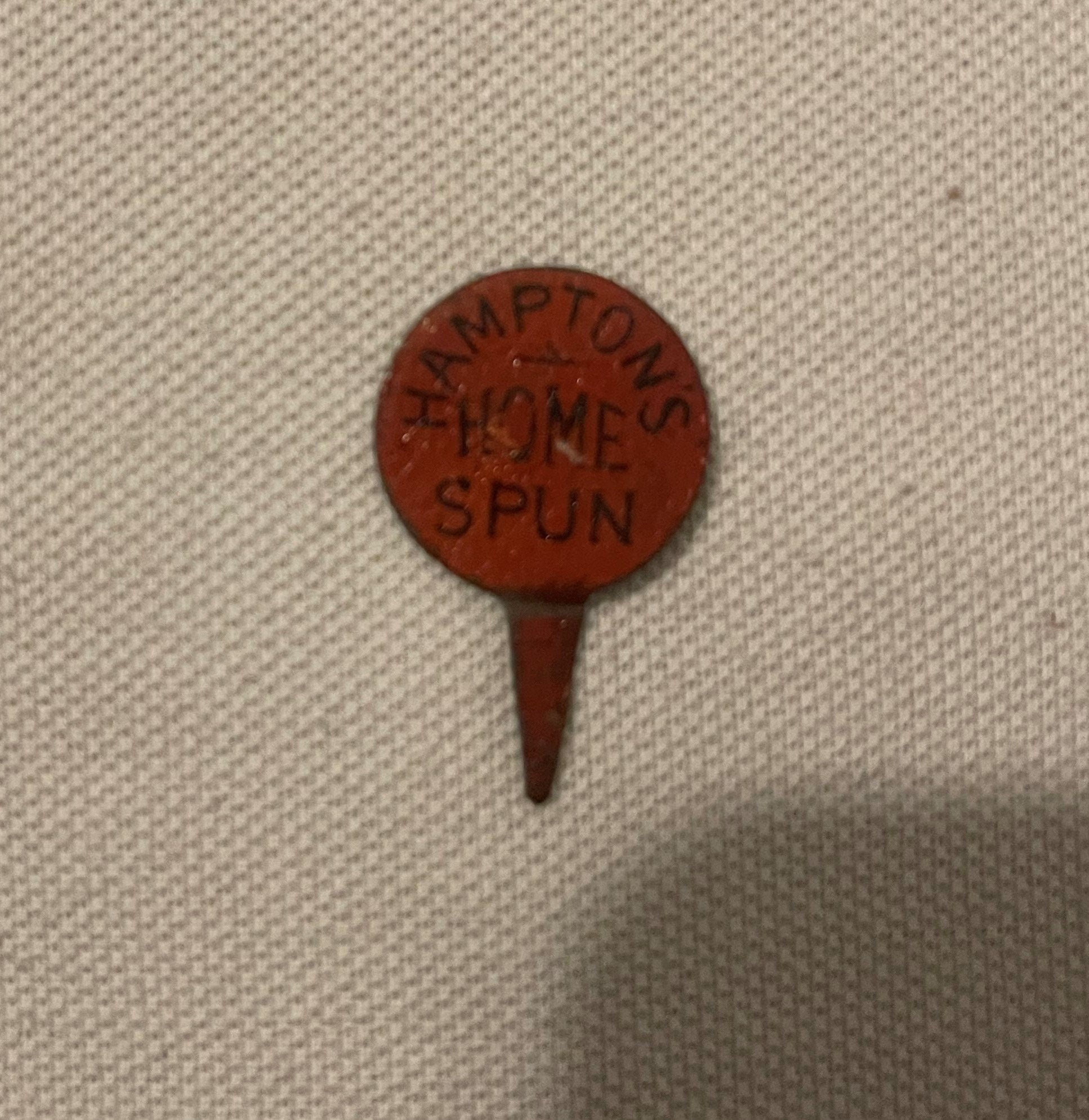 Red circular embossed tin advertising pin with "HAMPTONS HOME SPUN" text, metal pin backing, displayed on beige fabric for sc