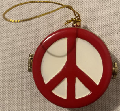 Red and white porcelain peace sign ornament with gold string and hinged metal clasp, displayed against beige fabric background.