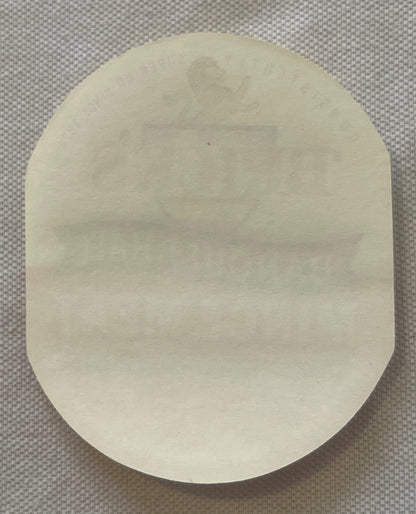 Cream-colored oval tin label with faint embossed text, photographed flat against gray textured background