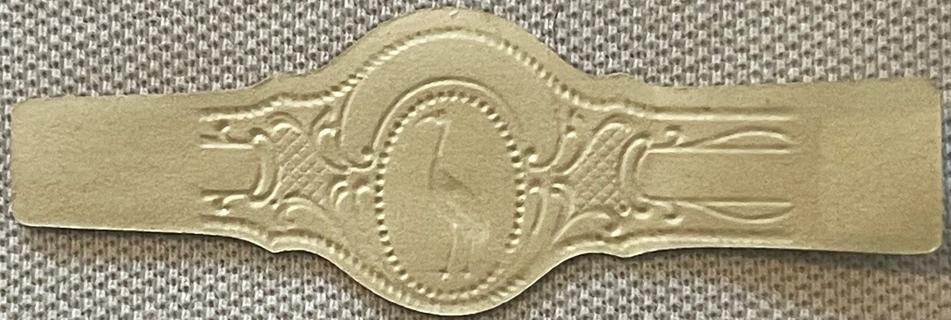 Gold embossed cigar band with ornate decorative borders and central oval portrait, displayed on textured fabric background