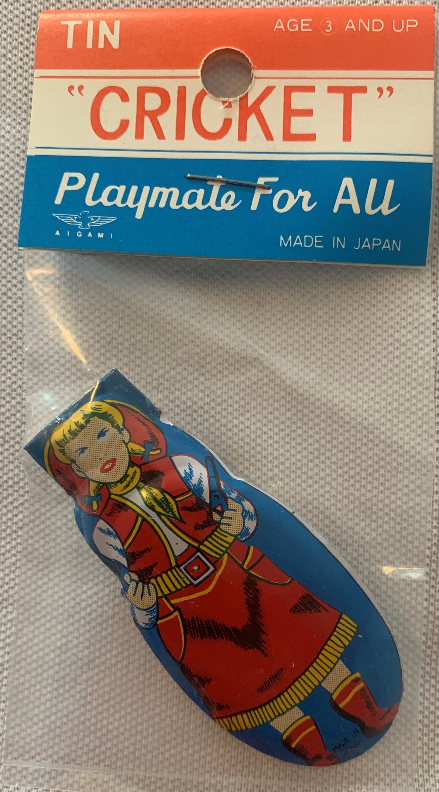 Packaged vintage tin cricket clicker with red, white and blue header and illustration of a girl in red traditional dress on b