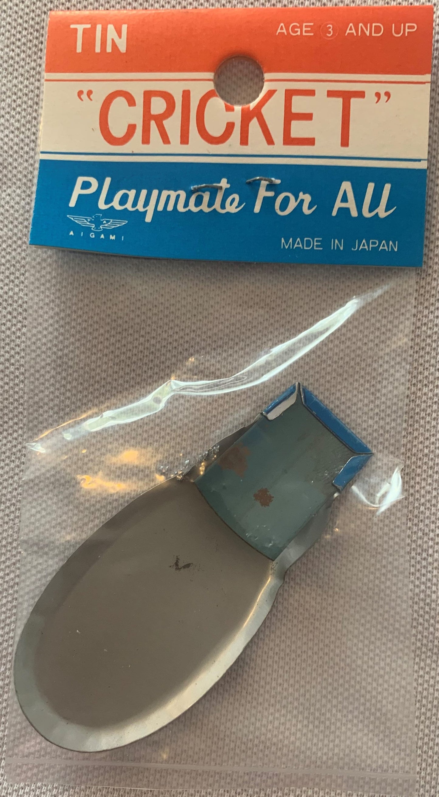Vintage tin cricket clicker with original packaging showing red, cream, and blue bands marked "Made in Japan," displayed with