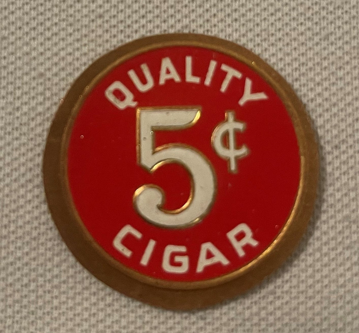 Round red and cream embossed metal token with "QUALITY 5¢ CIGAR" text and gold-colored border, photographed flat against beig