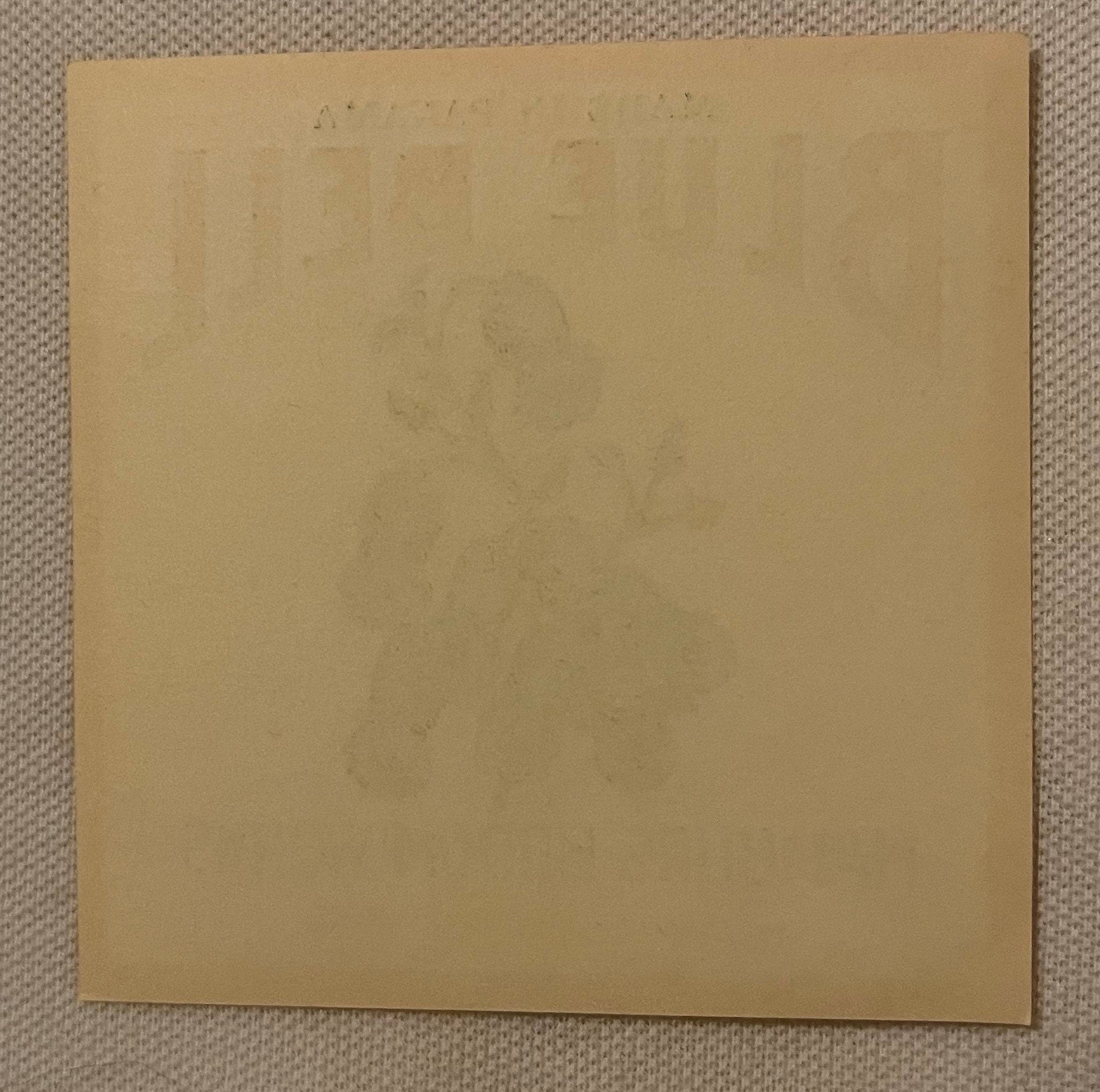 Blank tan cardboard sheet on textured beige fabric background, faint text visible at top edge