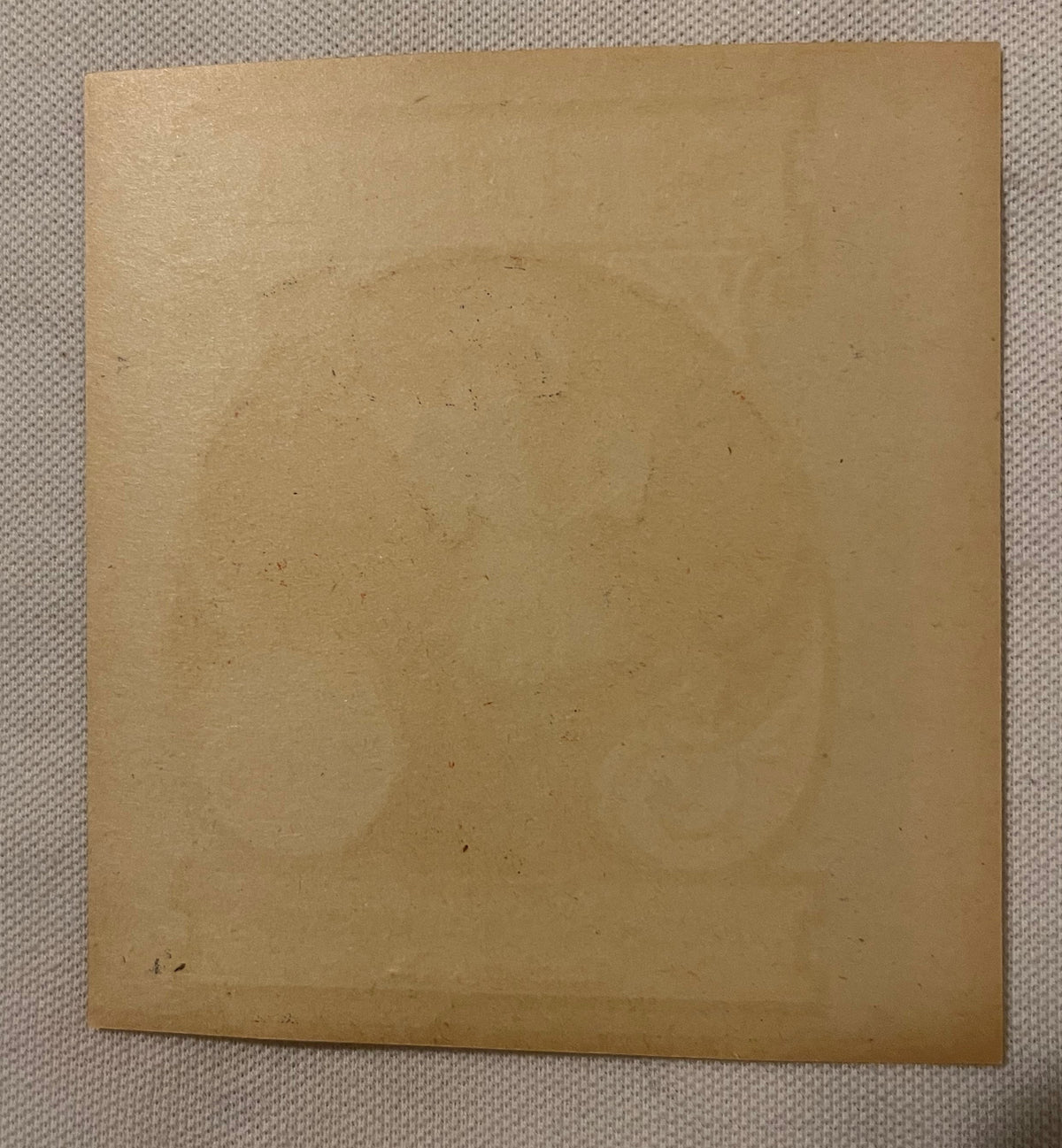 Tan cardboard label with scattered dark specks and wear marks, photographed flat against beige fabric surface