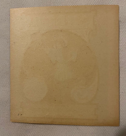 Tan cardboard label with scattered dark specks and wear marks, photographed flat against beige fabric surface