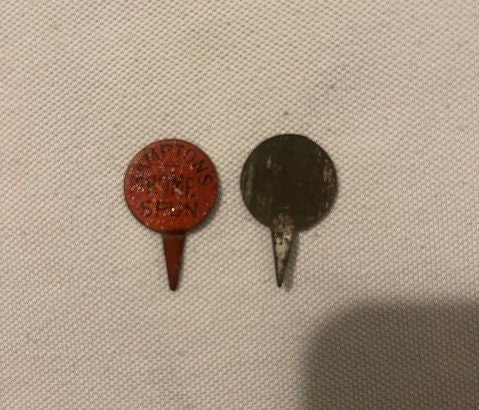 Two antique metal pin tacks on cream fabric, one red embossed and one dark, each approximately one inch diameter with pointed