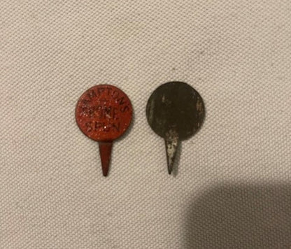 Two antique metal pin tacks on cream fabric, one red embossed and one dark, each approximately one inch diameter with pointed