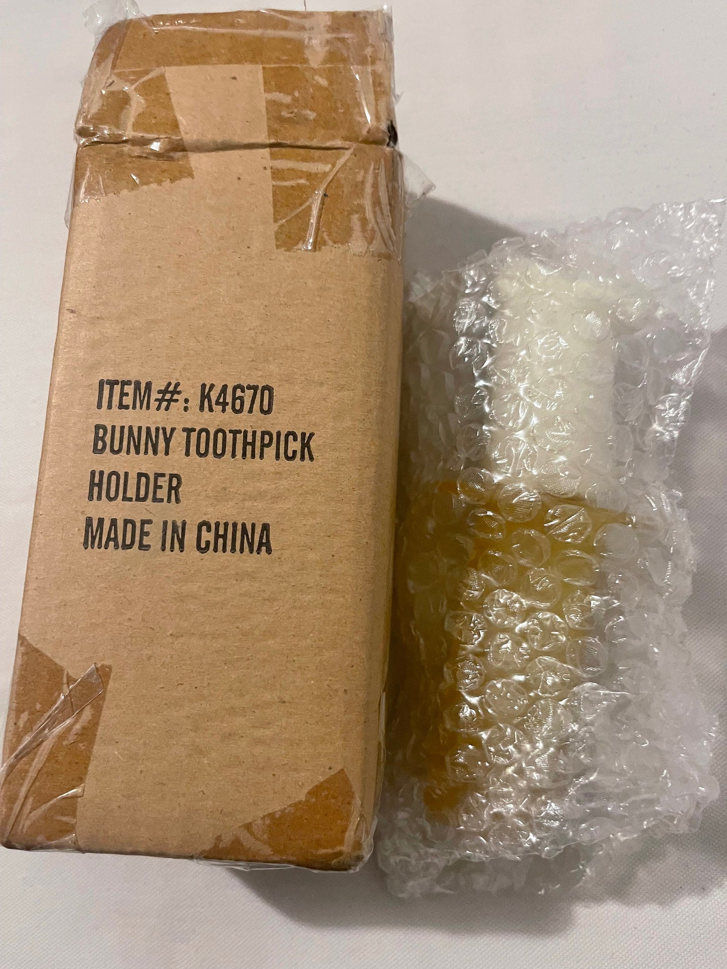 Cardboard box labeled "ITEM# K4670 BUNNY TOOTHPICK HOLDER MADE IN CHINA" next to plastic-wrapped amber glass rabbit figurine 
