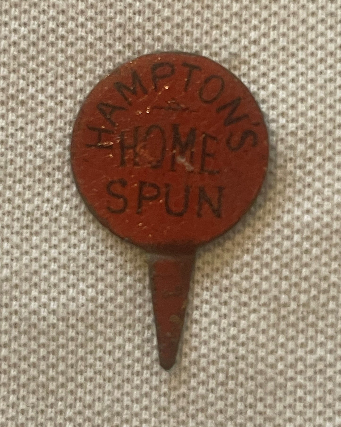 Circular red clay advertising pin with embossed text reading "HAMPTON'S HOME SPUN" displayed against cream fabric background