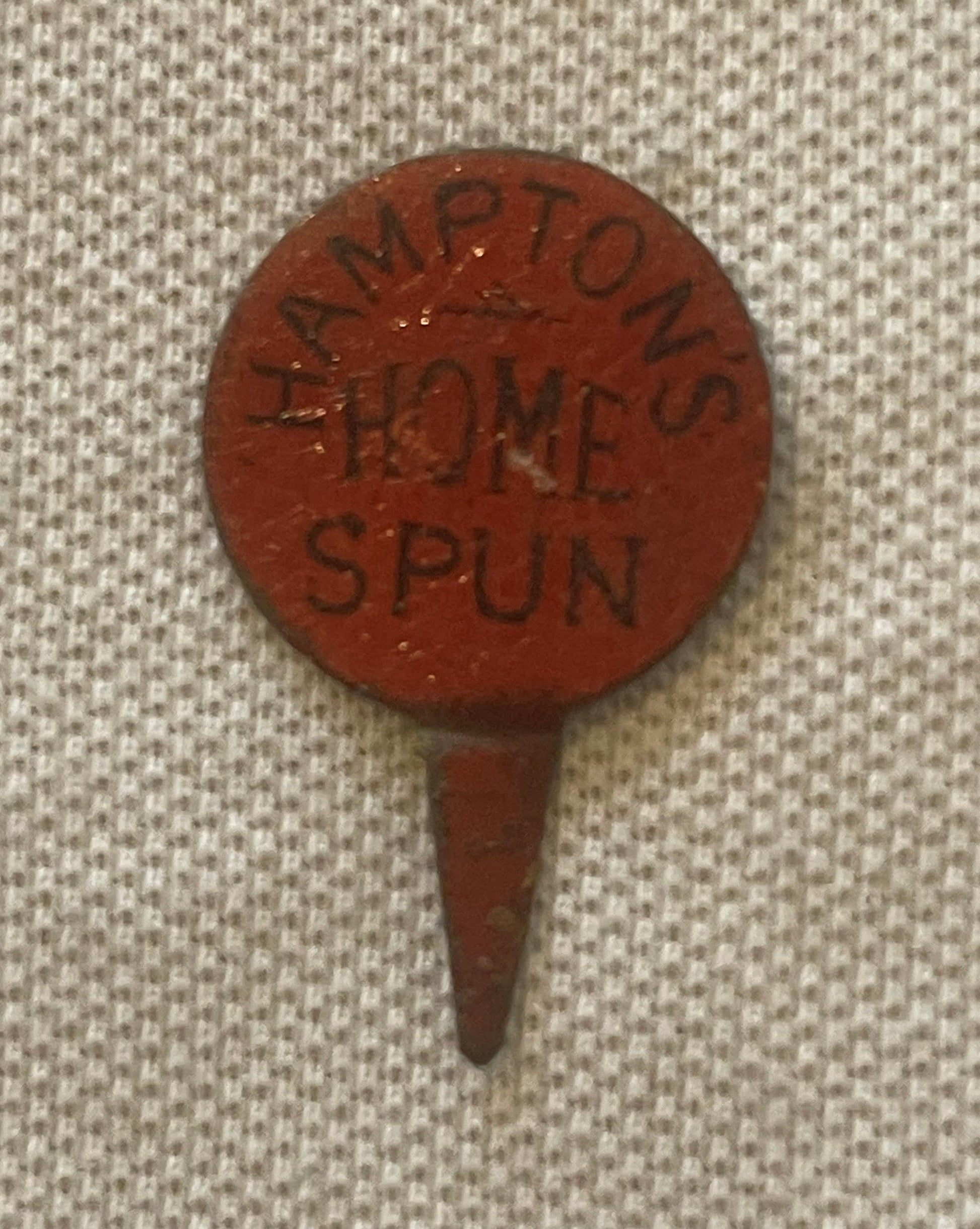 Circular red clay advertising pin with embossed text reading "HAMPTON'S HOME SPUN" displayed against cream fabric background