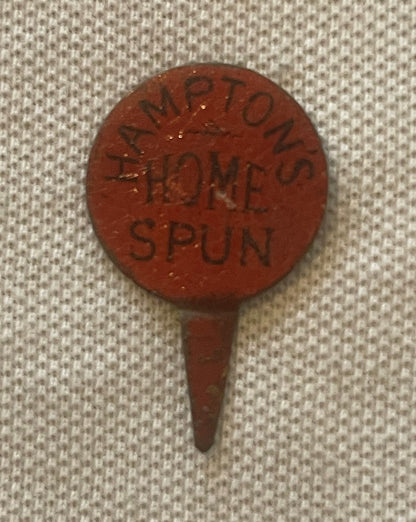 Circular red clay advertising pin with embossed text reading "HAMPTON'S HOME SPUN" displayed against cream fabric background