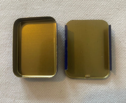 A FIXACO tin opened, its base and lid laid side by side, both empty with plain gold-toned interiors.