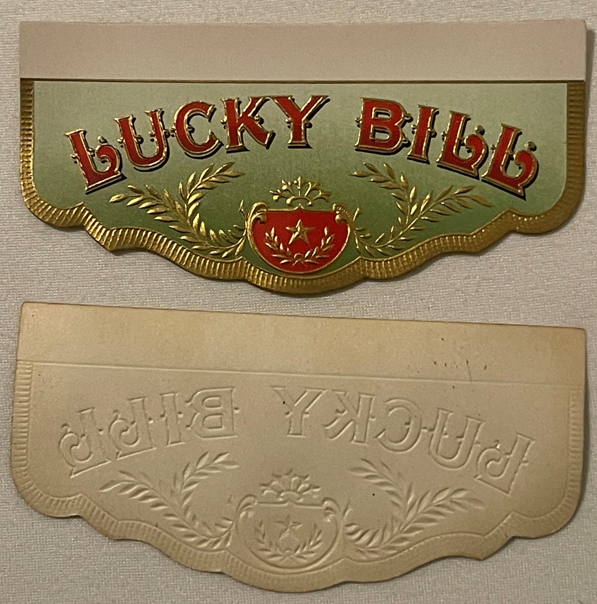 Two embossed cigar box label pieces with "Lucky Bill" in red and gold lettering, featuring decorative laurel wreaths and shie