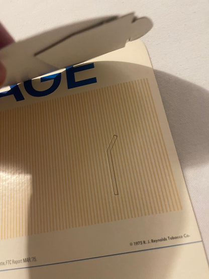 Close-up of cream-colored vintage cigarette display box with blue "GE" text visible at top, embossed vertical lines pattern, 
