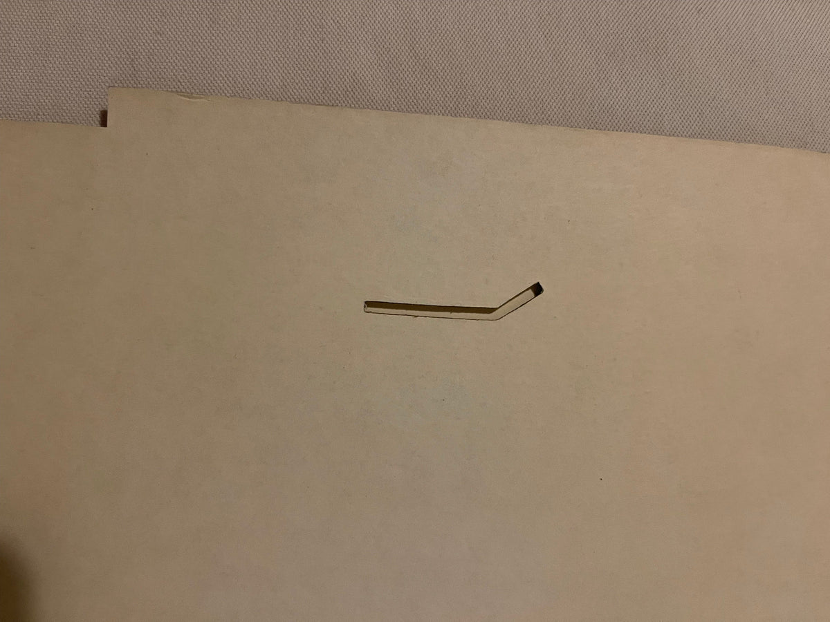 Bent metal wire bracket or support piece on cream-colored cardboard backing, photographed flat against textured gray surface