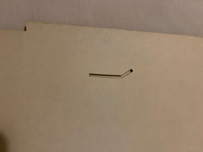 Bent metal wire bracket or support piece on cream-colored cardboard backing, photographed flat against textured gray surface