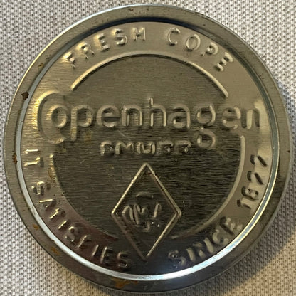 Circular brass tin lid embossed with "Copenhagen" and "It Satisfies Since 1822" text, displayed flat on gray fabric showing w