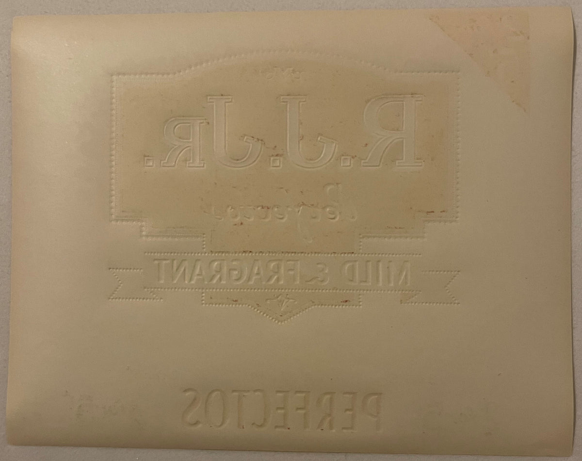 Reverse of the R.J. Jr. Perfectos label: blank cream paper with the blind-embossed mirror-image of the front panel - backward R.J.JR., Mild & Fragrant, and Perfectos lettering, with scattered ink offset marks, no color.