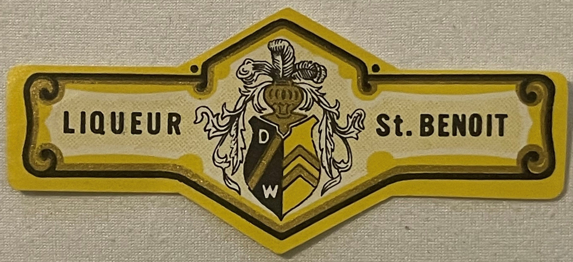 Vintage yellow and black heraldic neck label for St. Benoit liqueur with ornate coat of arms crest, mounted on textured beige
