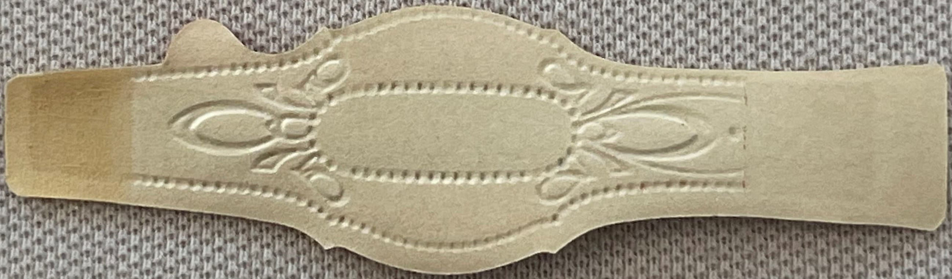 Cream-colored embossed cigar band with ornate floral scrollwork and central oval cartouche, displayed flat against textured f