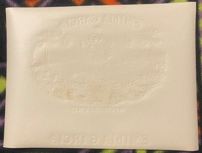 Cream-colored embossed cigar box label with ornamental border and text, photographed at an angle against a patterned fabric b