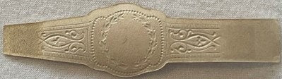 Embossed gold cigar band with ornamental side designs and blank central medallion, photographed flat against beige fabric