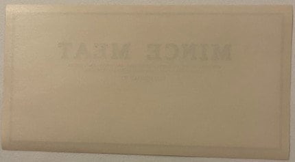 Back of the unprinted mince meat stock label: blank cream paper with no printing at all, showing only the front's MINCE MEAT lettering ghosting through the sheet in mirror image