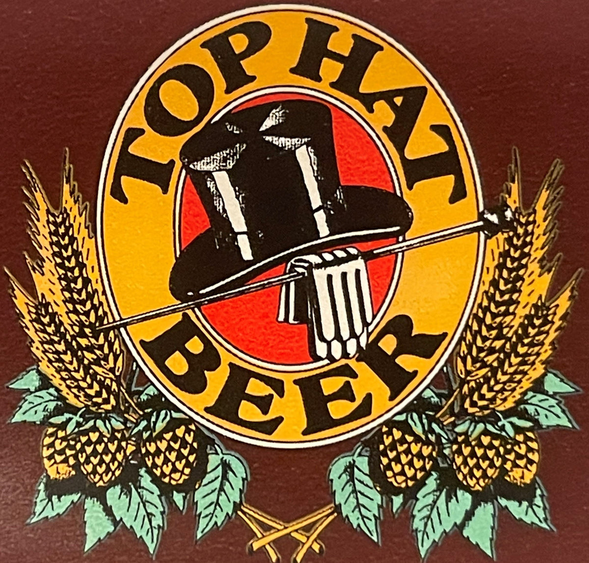 Close-up of the Top Hat Beer roundel: a black silk top hat on its side inside a red disc, a walking cane laid across the brim and a tankard in front, ringed in gold lettered TOP HAT above and BEER below, with barley sheaves and hop cones around it.