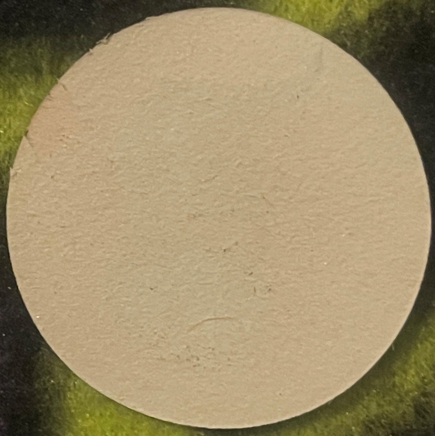 Cream-colored circular wine barrel label on green fabric background, blank face with subtle texture visible