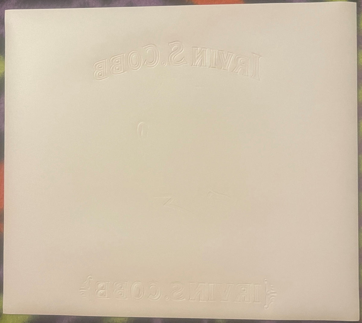 Cream-colored embossed cigar label with reversed text visible at top and bottom, photographed flat against multicolored backg