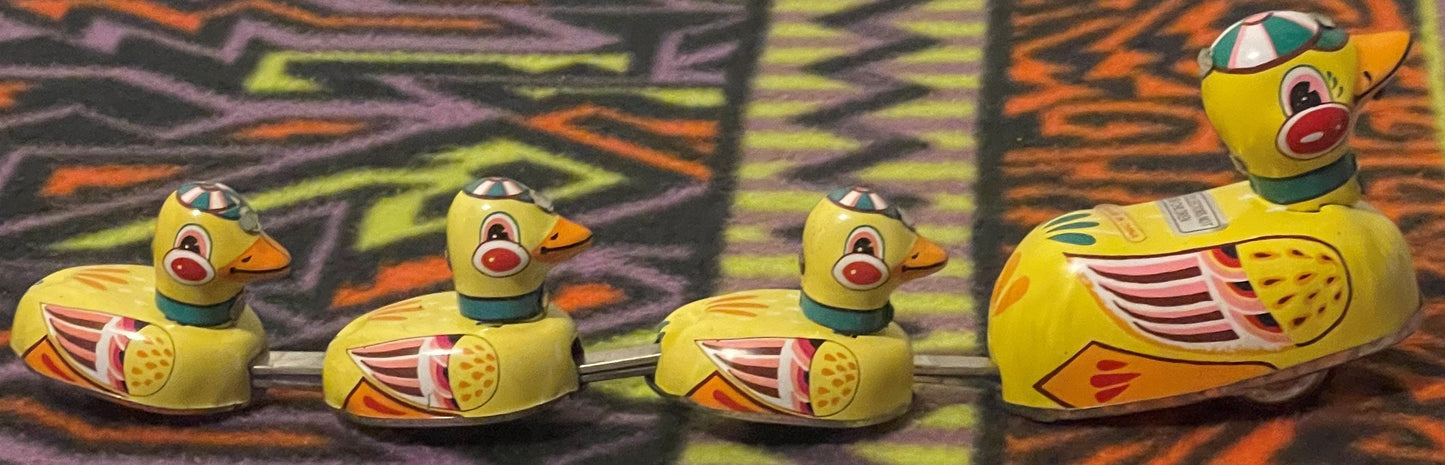 The mother duck and her three ducklings joined in a row by their metal linking rods, seen side on against patterned fabric.
