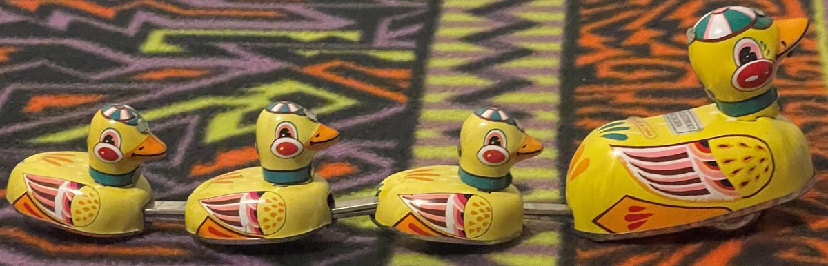 The mother duck and her three ducklings joined in a row by their metal linking rods, seen side on against patterned fabric.