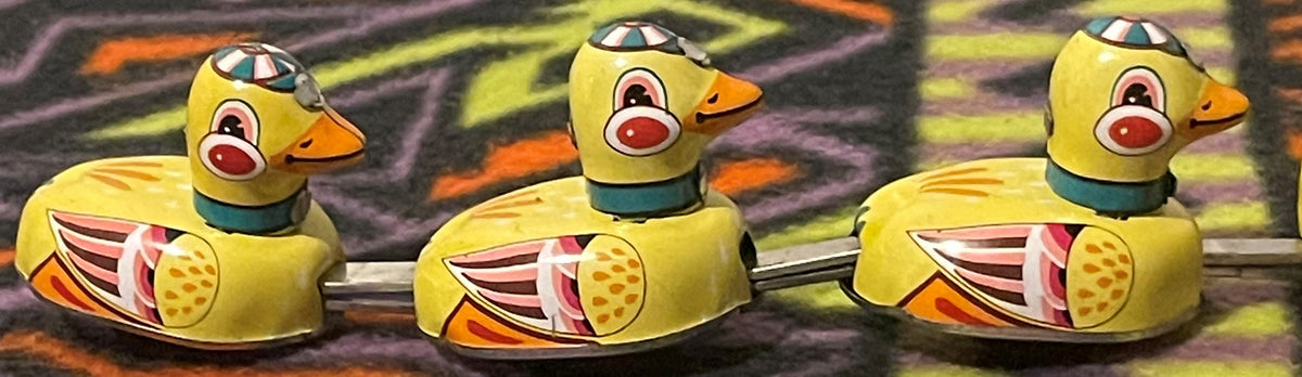 The three small ducklings alone in a row, linked by their metal rods, each with a teal collar and painted racing cap.