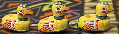 The three small ducklings alone in a row, linked by their metal rods, each with a teal collar and painted racing cap.