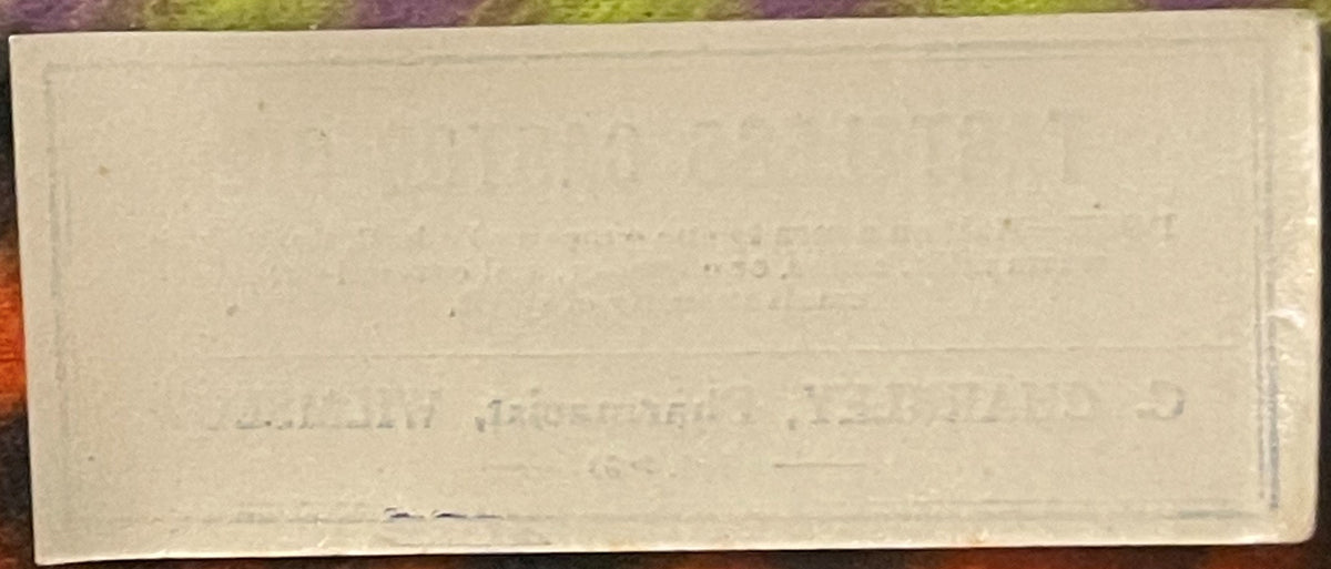 Cream-colored antique label with printed text, displayed flat against a wooden surface with colorful fabric visible at