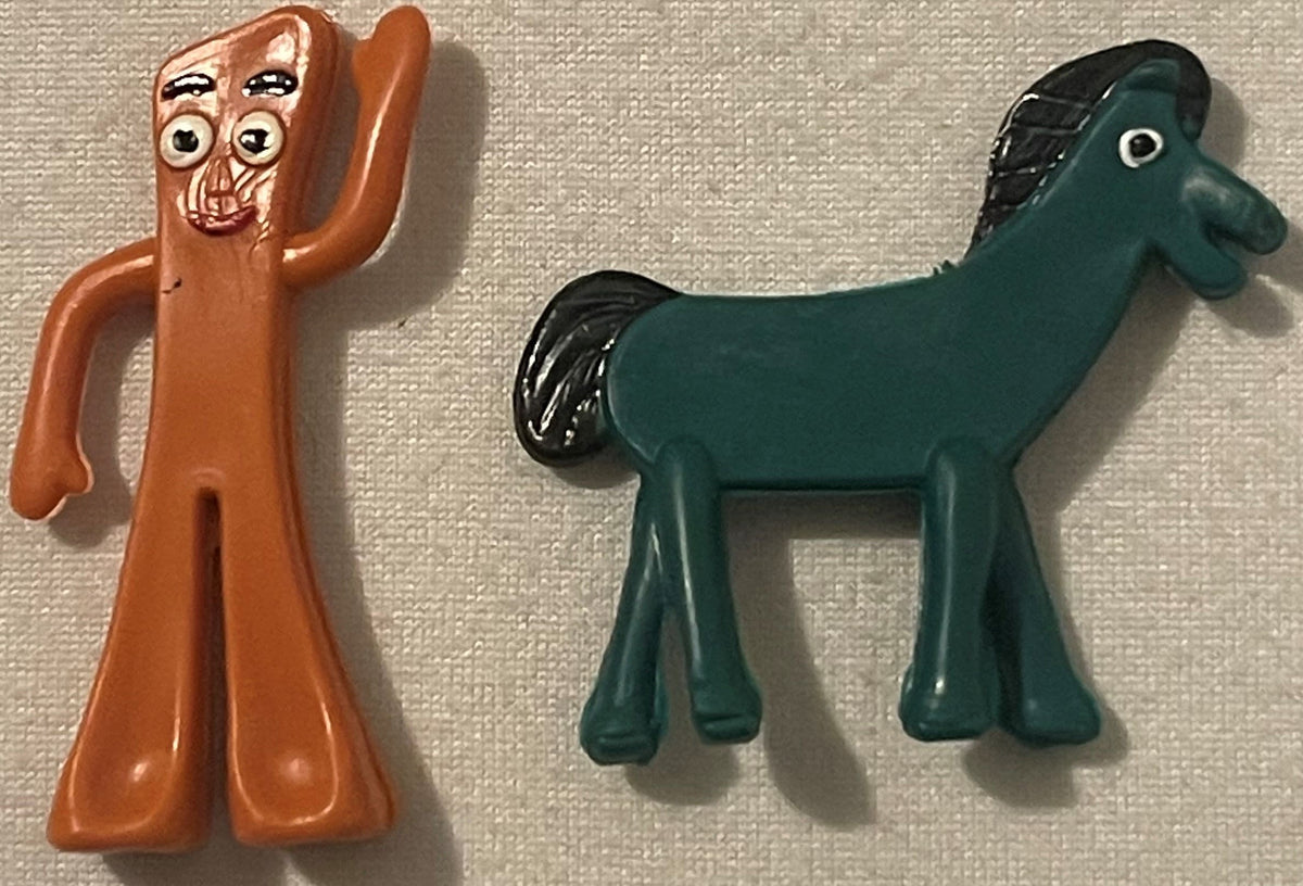 Close view of an orange-brown Gumby with hand-painted eyes and a small red smile beside a teal Pokey horse with a black molded mane, both resting on woven fabric.
