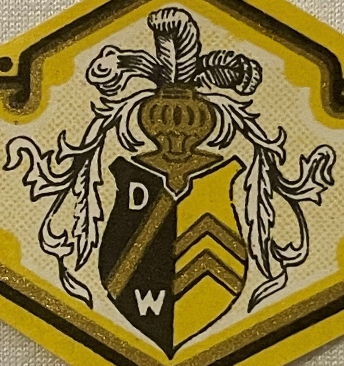 Antique heraldic coat of arms label with yellow and black shield, ornate leafy wings, and letters D and W on cream background