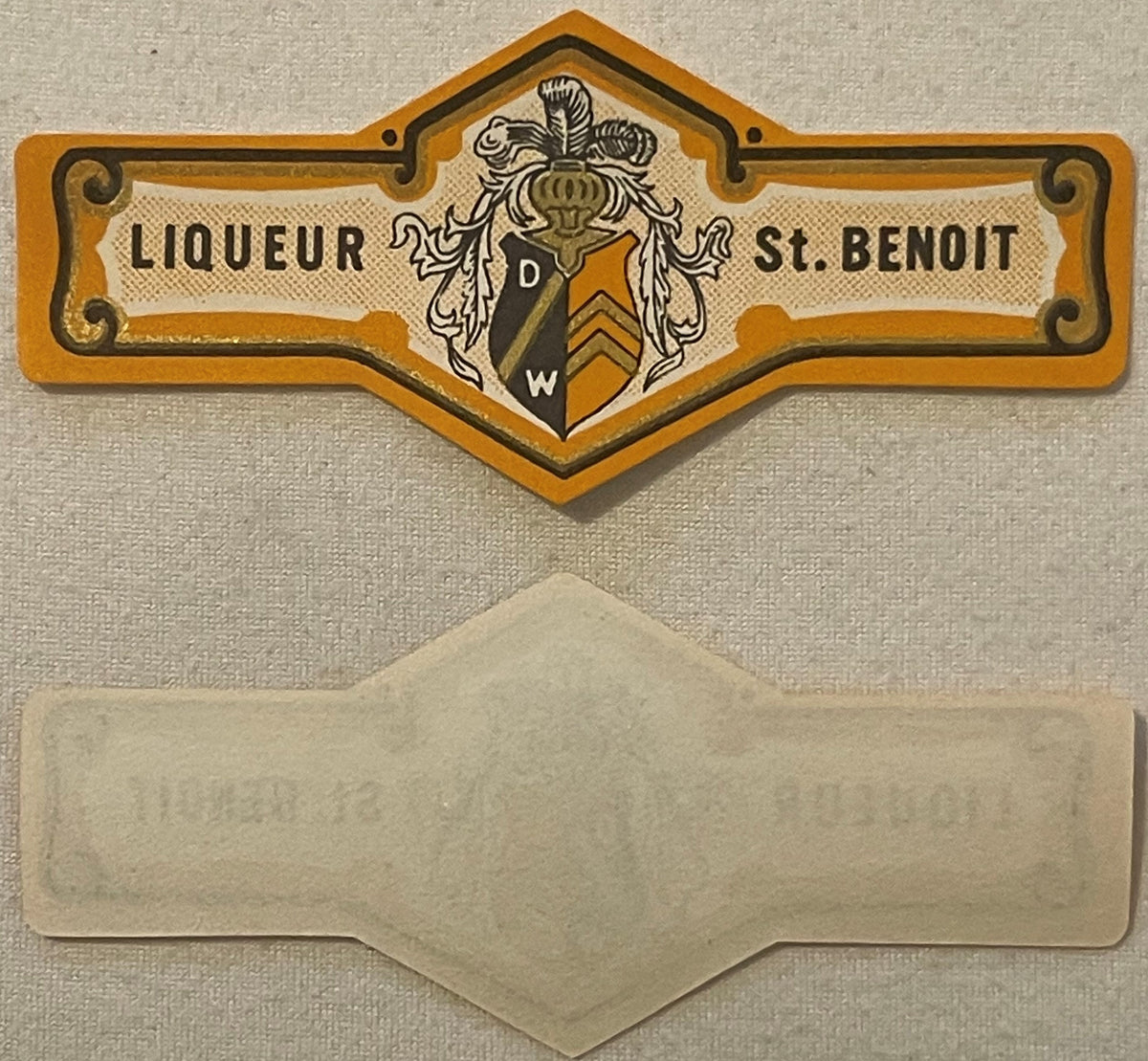 Vintage Liqueur St. Benoit bottle neck label with orange heraldic crest and blank cream-colored band below, displayed flat ag