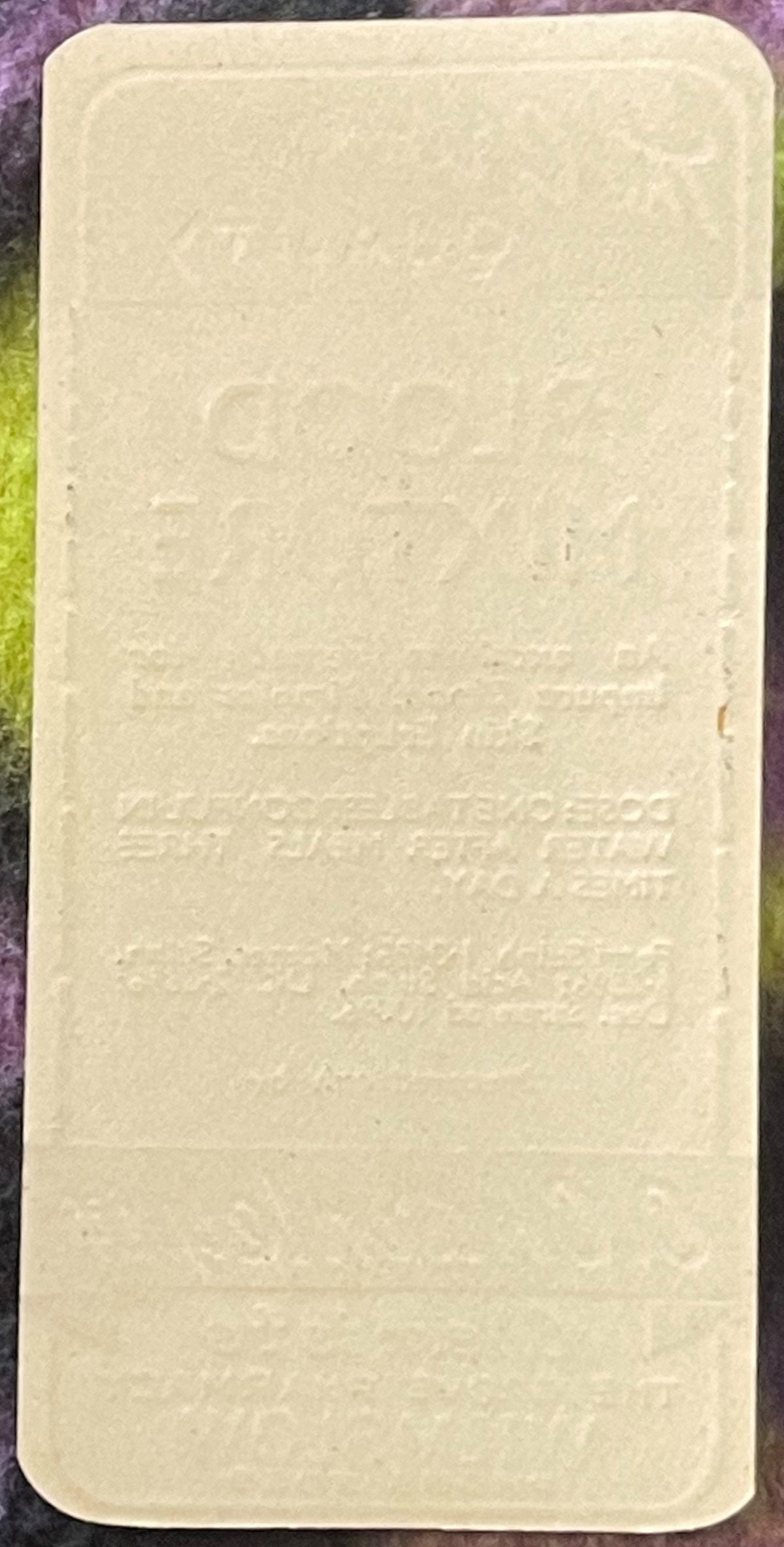 Cream-colored rectangular pharmacy label with embossed text, photographed flat against purple and green fabric background