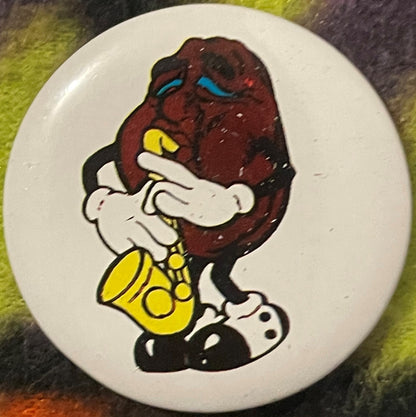 Close-up of one pinback button featuring a cartoon raisin character with dark brown head, blue eyes, yellow nose, white glove