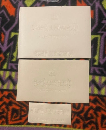 All three Cornwall Arms labels arranged vertically - white, orange wood-grain, and the small label - on a colorful patterned surface for scale