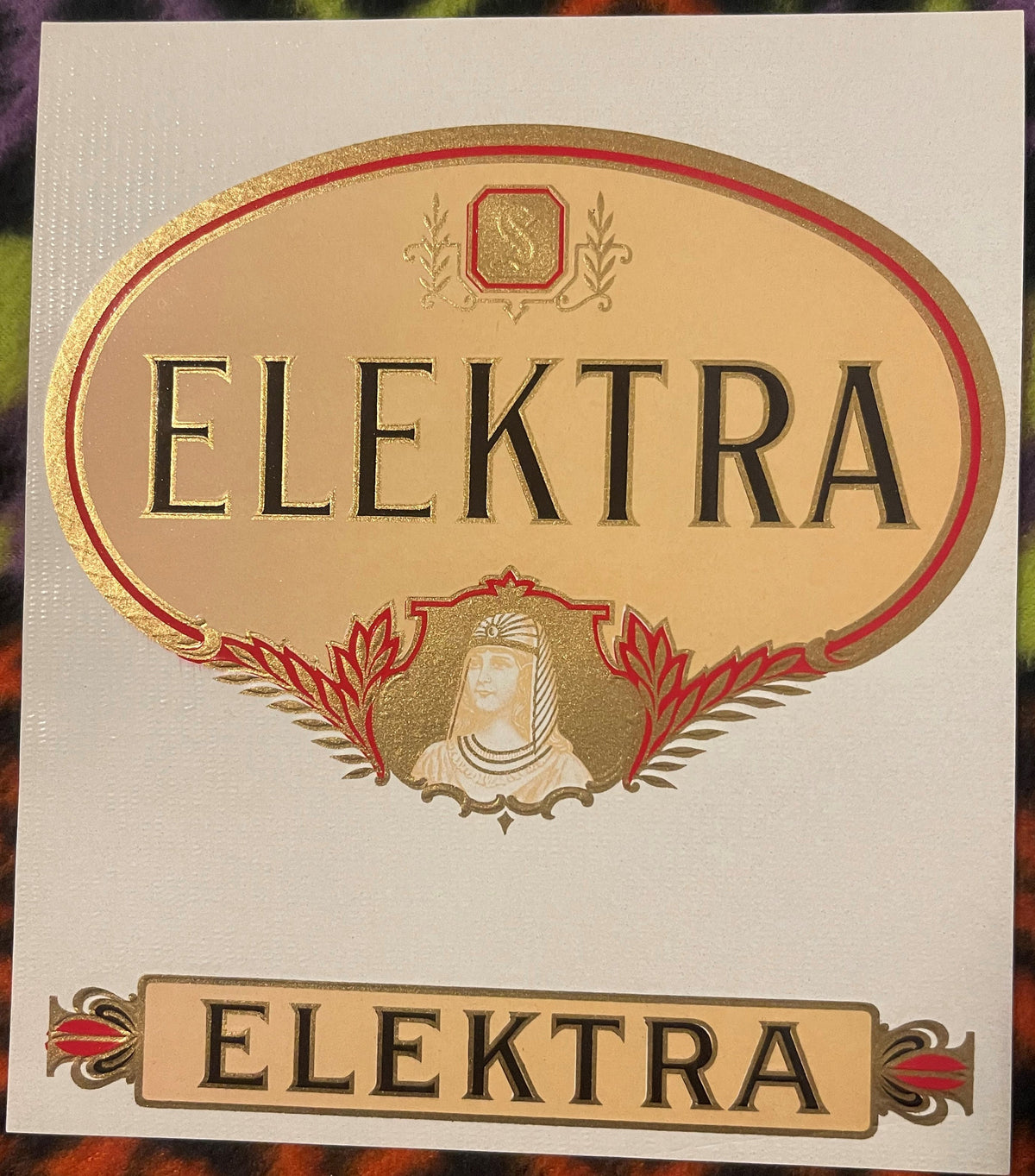 Elektra gold embossed cigar label: ELEKTRA in a large gilt oval with red borders, a script monogram in an octagon at the top, a woman in a striped Egyptian-style headdress below, and a matching ELEKTRA title strip