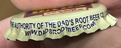 Cream-colored bottle cap held in a person's hand, printed with "The Authority of the Dad's Root Beer Co." and website text in