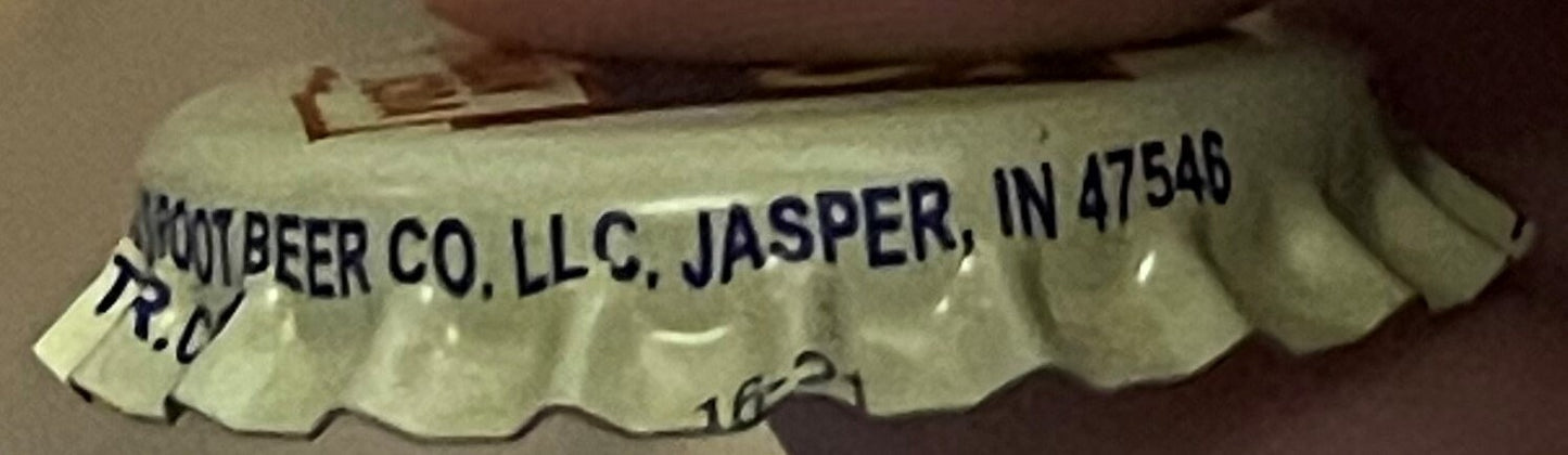 Vintage cream soda bottle cap printed "Kumbeer Co. LLC, Jasper, IN 47546" held at an angle against a brown surface