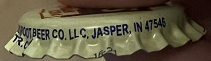 Vintage cream soda bottle cap printed "Kumbeer Co. LLC, Jasper, IN 47546" held at an angle against a brown surface